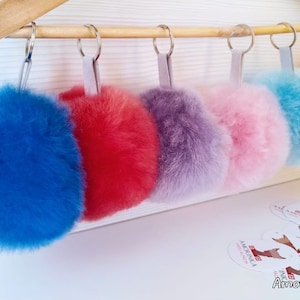 May include: Six colorful fluffy pom-poms on keychains, hanging from a wooden hanger. The pom-poms are blue, red, purple, pink, light blue, and yellow.