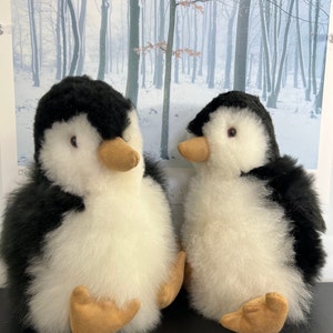 May include: Two plush penguin toys, one black and white and one smaller, with a white body and black wings, are sitting on a black surface. The penguins have brown beaks and black eyes.