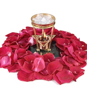 May include: A lit candle in a brass oil burner with a gold-colored top, filled with white crystals, sits on a dark green surface, surrounded by red rose petals. The burner has a star-shaped base.