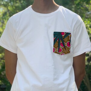 以下が含まれることがあります： カラフルなアフリカンプリントのデザインが施されたポケットが付いた白いTシャツ。プリントにはピンク、ブラウン、イエロー、グリーンの色が含まれています。
