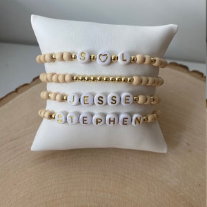 May include: Four wooden bead bracelets with white letter beads spelling out the names "SOL", "JESSE", and "STEPHEN". The bracelets are stacked on a white pillow.