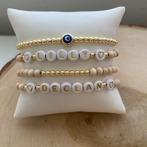 May include: Four bracelets stacked on a white display stand. The top bracelet is a gold chain with a blue evil eye charm. The next bracelet is white with gold letters spelling "LILLY" with a heart on each end. The third bracelet is a natural wood bead bracelet. The bottom bracelet is white with gold letters spelling "DECLAN" with a heart on each end.