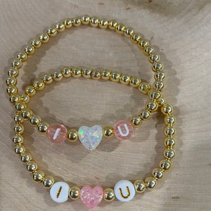 May include: Two gold beaded bracelets with the letters "I" and "U" separated by a pink heart charm. The charms are made of plastic and have a glittery finish.