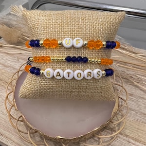 May include: Three beaded bracelets with gold accents. The top bracelet has orange and blue beads with the letters "UF" in white. The middle bracelet has orange and blue beads. The bottom bracelet has blue beads with the letters "GATORS" in white.