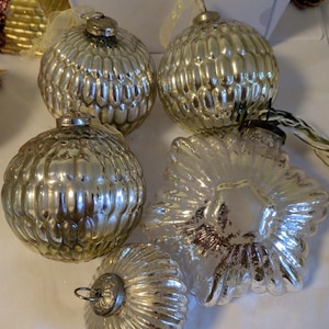 May include: A collection of vintage Christmas ornaments in various shapes. The ornaments are silver and gold, with ribbed and floral designs. Some have ribbons for hanging. The ornaments are arranged on a white surface.