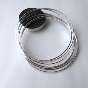 May include: Several silver bangles, each a thin, shiny circle, are displayed on a white background. The bangles reflect light. A small, dark grey, circular object is partially visible in the background.