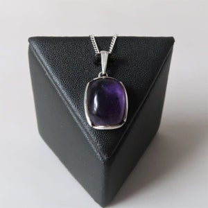 May include: A square, purple amethyst pendant with a silver-coloured frame and chain. The gemstone is a deep, translucent purple. The pendant is displayed on a black, triangular stand against a white background.