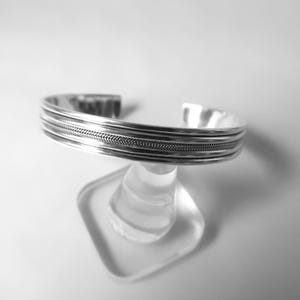 May include: A silver cuff bracelet with a textured design. The bracelet features multiple parallel bands, with a central band showcasing a woven pattern. The open-ended design allows for easy wear. The jewelry is displayed on a clear stand.