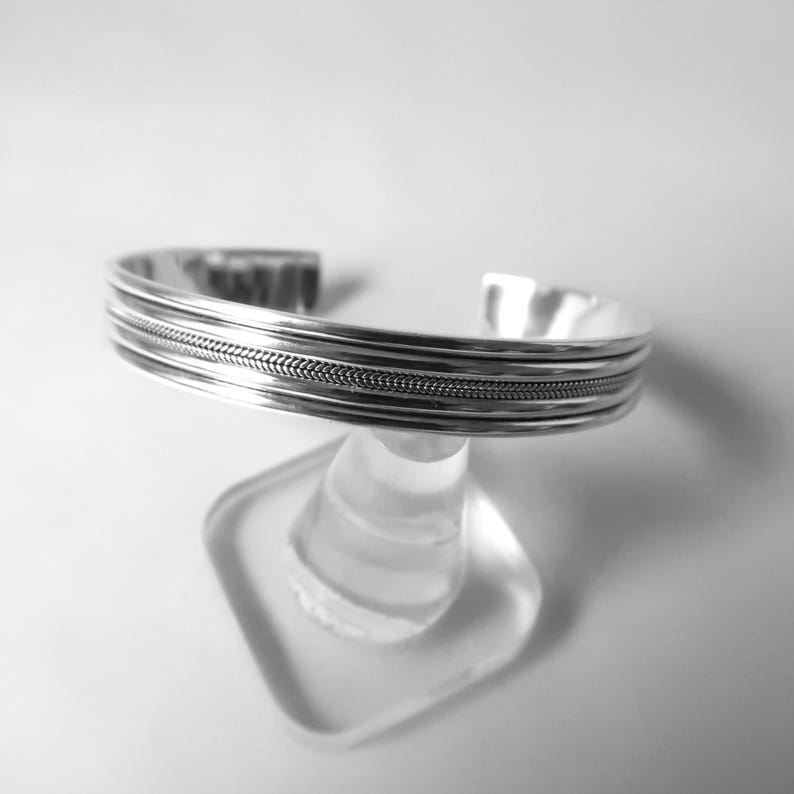 May include: A silver cuff bracelet with a textured design. The bracelet features multiple parallel bands, with a central band showcasing a woven pattern. The open-ended design allows for easy wear. The jewelry is displayed on a clear stand.