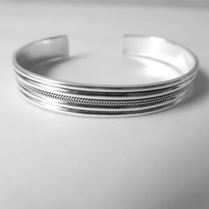 May include: A silver cuff bracelet with a textured, braided design in the center. The bracelet has a simple, open design and is made of a shiny, reflective metal. The bracelet is a piece of jewelry.