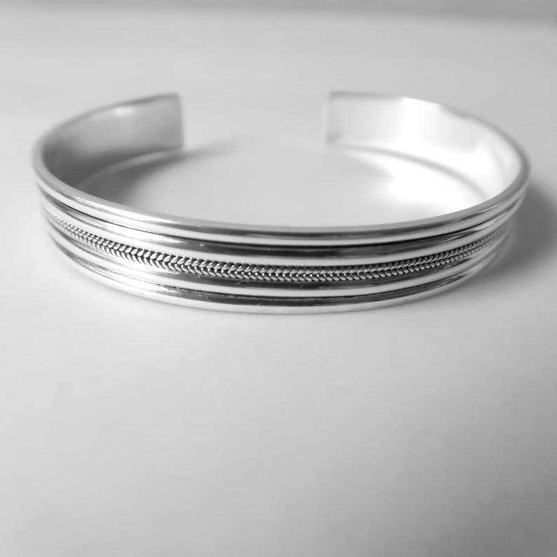 May include: A silver cuff bracelet with a textured, braided design in the center. The bracelet has a simple, open design and is made of a shiny, reflective metal. The bracelet is a piece of jewelry.