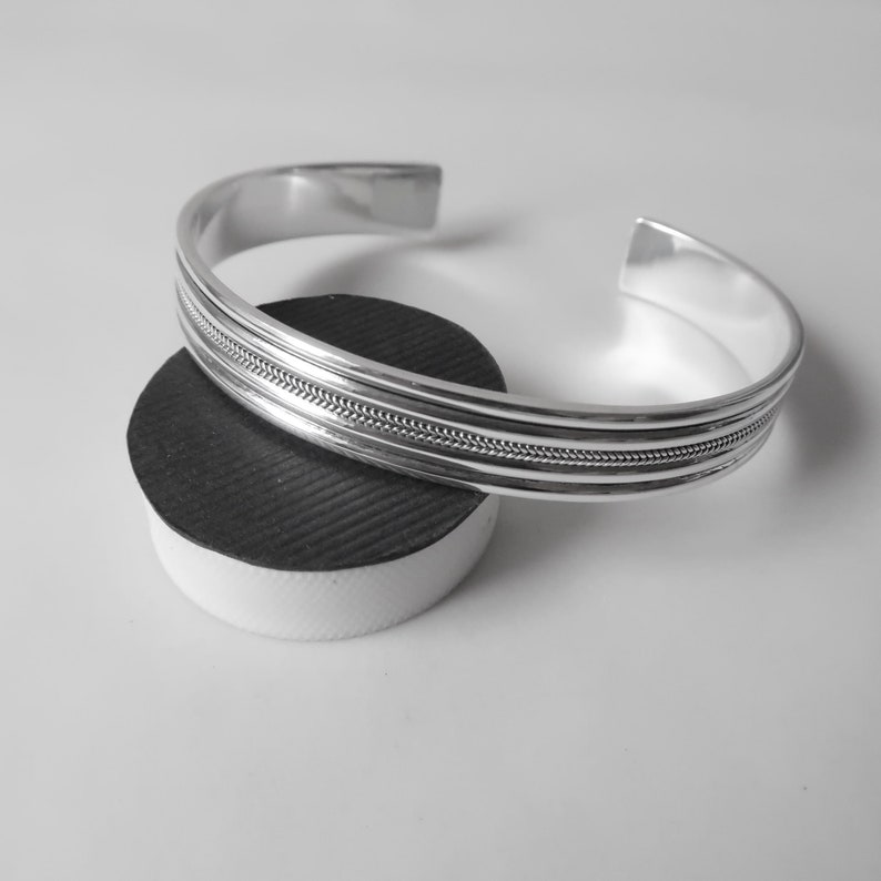 May include: A silver cuff bracelet with a textured band. The open-ended bracelet is displayed on a black and white cylindrical object. The bracelet has a polished finish and a simple, elegant design.