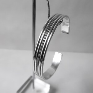 May include: A silver cuff bracelet with a braided center design. The bracelet has a polished finish and is displayed on a silver stand. The design features three parallel bands with a textured, woven pattern in the center.