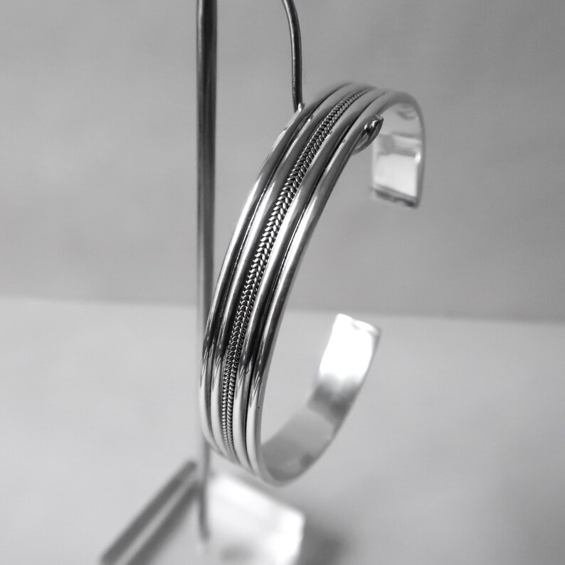 May include: A silver cuff bracelet with a braided center design. The bracelet has a polished finish and is displayed on a silver stand. The design features three parallel bands with a textured, woven pattern in the center.