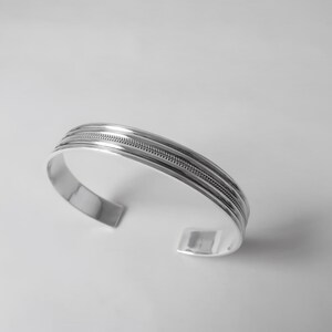 May include: A silver cuff bracelet with a polished finish. The bracelet features three parallel bands, with a textured, braided design in the center band. The open-ended design allows for easy wear.