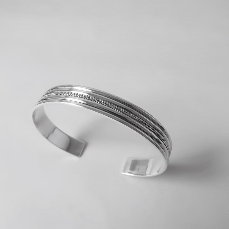 May include: A silver cuff bracelet with a polished finish. The bracelet features three parallel bands, with a textured, braided design in the center band. The open-ended design allows for easy wear.