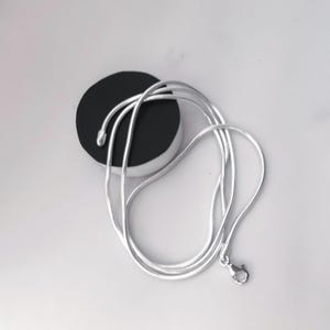 May include: A silver snake chain necklace with a lobster clasp, coiled on a white surface. The necklace is draped over a black and white circular object.