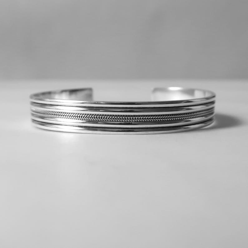 May include: A silver cuff bracelet with a textured, woven design in the center. The bracelet has a simple, elegant design with three parallel lines above and below the textured section. The bracelet is shown on a white surface.