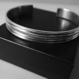 May include: A silver cuff bracelet with a textured, braided design in the center. The bracelet is displayed on a black box, creating a contrast between the shiny metal and the dark background. The bracelet has a simple, elegant design.