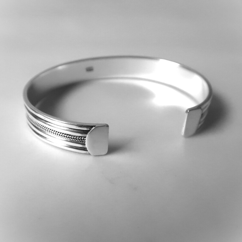 May include: A silver cuff bracelet with a textured design. The open-ended bracelet features a series of parallel lines and a central section with a woven pattern. The bracelet is made of a shiny, reflective metal.