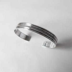 May include: A silver cuff bracelet with a textured design. The bracelet has a series of parallel bands, with a central band featuring a woven pattern. The open-ended design allows for easy wear.