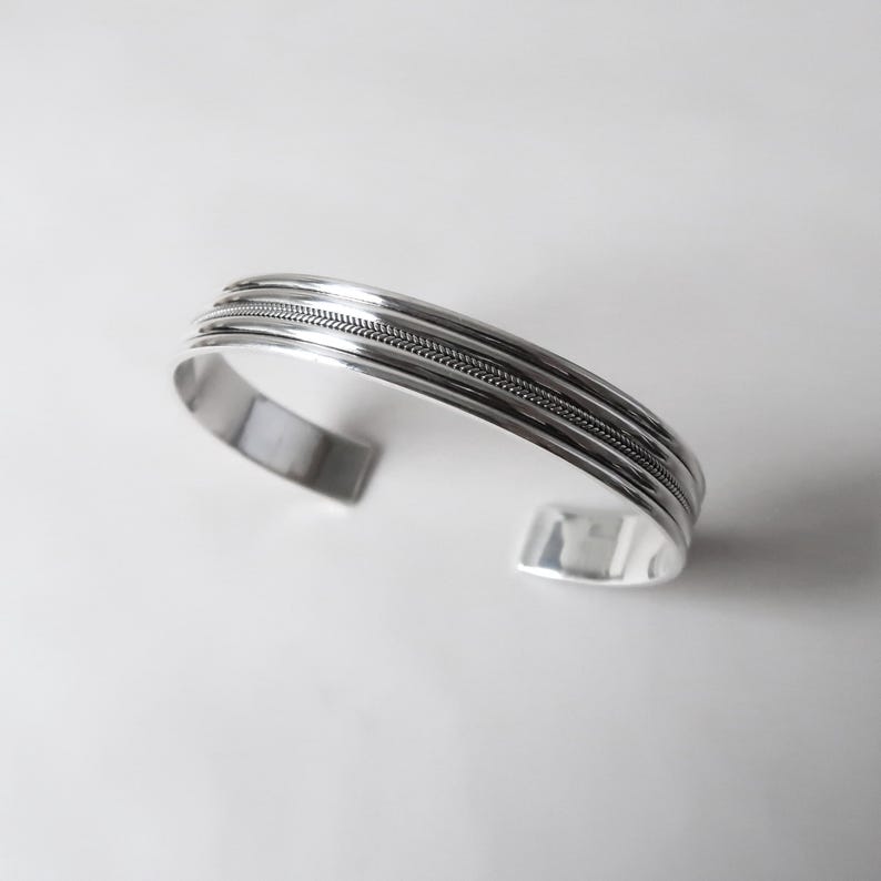 May include: A silver cuff bracelet with a textured design. The bracelet has a series of parallel bands, with a central band featuring a woven pattern. The open-ended design allows for easy wear.