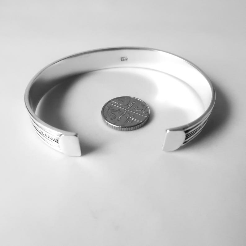 May include: A silver cuff bracelet with an open design, featuring a decorative pattern near the ends. A coin is placed inside the bracelet. The bracelet has a polished finish and a minimalist aesthetic, suitable for everyday wear.