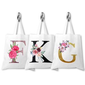 May include: Three white canvas tote bags with floral designs and letters F, K, and G. The F is pink and gold, the K is black, and the G is gold.