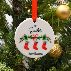 May include: White ceramic Christmas ornament with a red ribbon. The ornament features a green holly wreath with red berries and three red stockings with the names "Daddy", "Mommy", and "Sophia" written on them. The text "The Smith's" and "Merry Christmas" is also printed on the ornament.