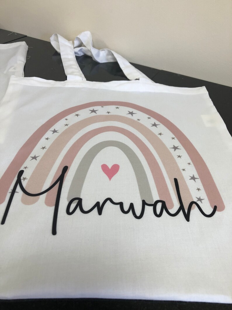 K&ouml;nnte beinhalten: Wei&szlig;e Einkaufstasche mit einem rosafarbenen und grauen Regenbogenmuster, Sternen und einem rosa Herzen. Der Name "Marwah" ist in schwarzer Kursivschrift unter dem Regenbogen geschrieben.