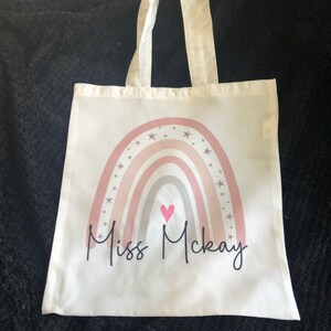 K&ouml;nnte beinhalten: Eine wei&szlig;e Stofftasche mit einem Regenbogen-Design und Sternen. Der Regenbogen ist in den Farben Rosa, Grau und Wei&szlig; gehalten. In der Mitte des Regenbogens befindet sich ein kleines pinkfarbenes Herz. Der Text "Miss McKay" ist unter dem Regenbogen aufgedruckt.