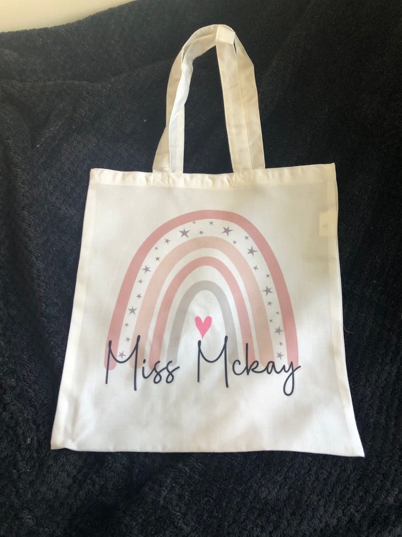 K&ouml;nnte beinhalten: Eine wei&szlig;e Stofftasche mit einem Regenbogen-Design und Sternen. Der Regenbogen ist in den Farben Rosa, Grau und Wei&szlig; gehalten. In der Mitte des Regenbogens befindet sich ein kleines pinkfarbenes Herz. Der Text "Miss McKay" ist unter dem Regenbogen aufgedruckt.