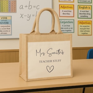 K&ouml;nnte beinhalten: Eine beigefarbene Jutetasche mit einer wei&szlig;en Vordertasche. Die Tasche tr&auml;gt den schwarzen Schriftzug "Mrs Smith's TEACHER STUFF" sowie ein Herzsymbol. Die Tasche hat zwei beige Griffe.