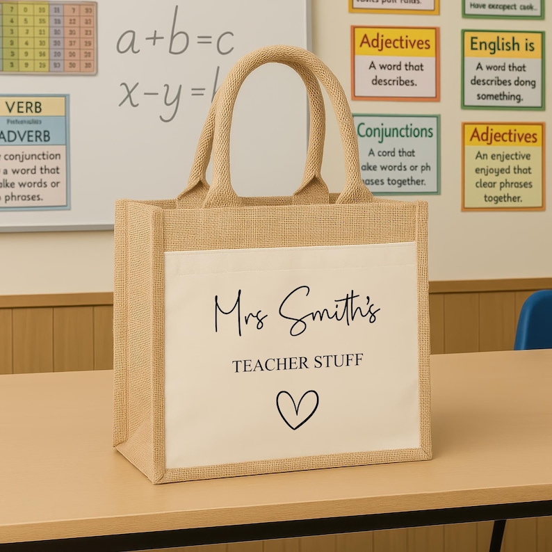 K&ouml;nnte beinhalten: Eine beigefarbene Jutetasche mit einer wei&szlig;en Vordertasche. Die Tasche tr&auml;gt den schwarzen Schriftzug "Mrs Smith's TEACHER STUFF" sowie ein Herzsymbol. Die Tasche hat zwei beige Griffe.