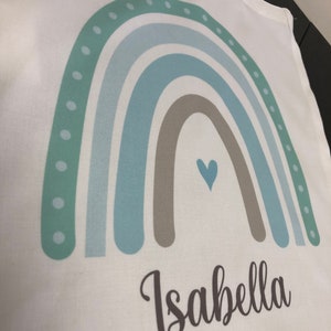 K&ouml;nnte beinhalten: Eine wei&szlig;e Tragetasche mit einem Regenbogen-Design in Gr&uuml;n-, Blau- und Braunt&ouml;nen. Der Regenbogen hat ein kleines Herz in der Mitte und der Name "Isabella" ist darunter gedruckt.