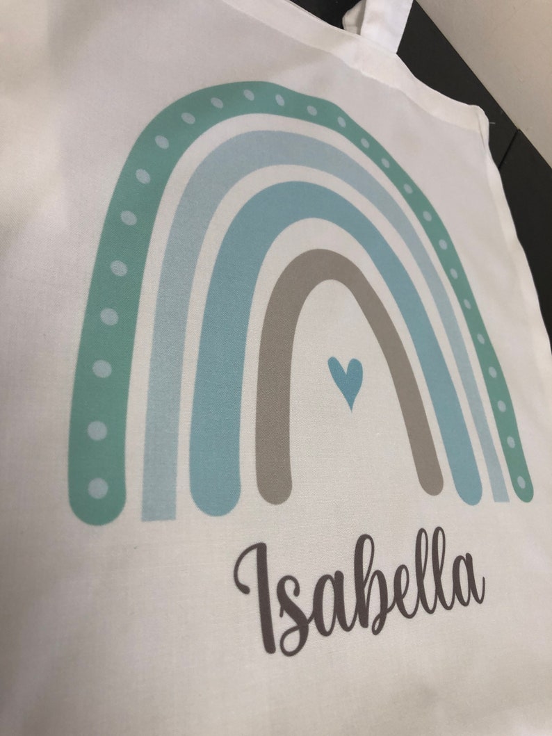 K&ouml;nnte beinhalten: Eine wei&szlig;e Tragetasche mit einem Regenbogen-Design in Gr&uuml;n-, Blau- und Braunt&ouml;nen. Der Regenbogen hat ein kleines Herz in der Mitte und der Name "Isabella" ist darunter gedruckt.