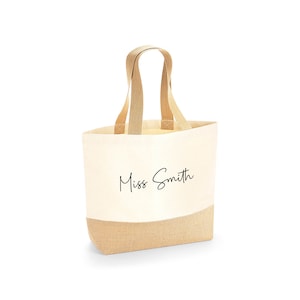 K&ouml;nnte beinhalten: Eine beigefarbene Canvas-Tragetasche mit einem hellbraunen Boden und passenden Griffen. Der Name "Miss Smith" ist in schwarzer Schrift auf der Vorderseite aufgedruckt. Die Tasche ist zum Tragen von Gegenst&auml;nden gedacht.