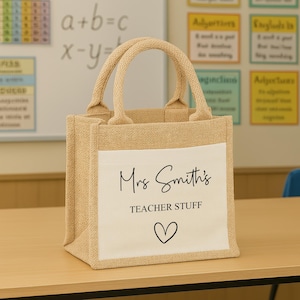 K&ouml;nnte beinhalten: Beige Jute-Tragetasche mit wei&szlig;er Fronttasche. Die Tasche tr&auml;gt den Text "Mrs Smith's TEACHER STUFF" und ein schwarzes Herzsymbol. Die Tasche hat zwei beige Griffe und steht auf einer Holzoberfl&auml;che.