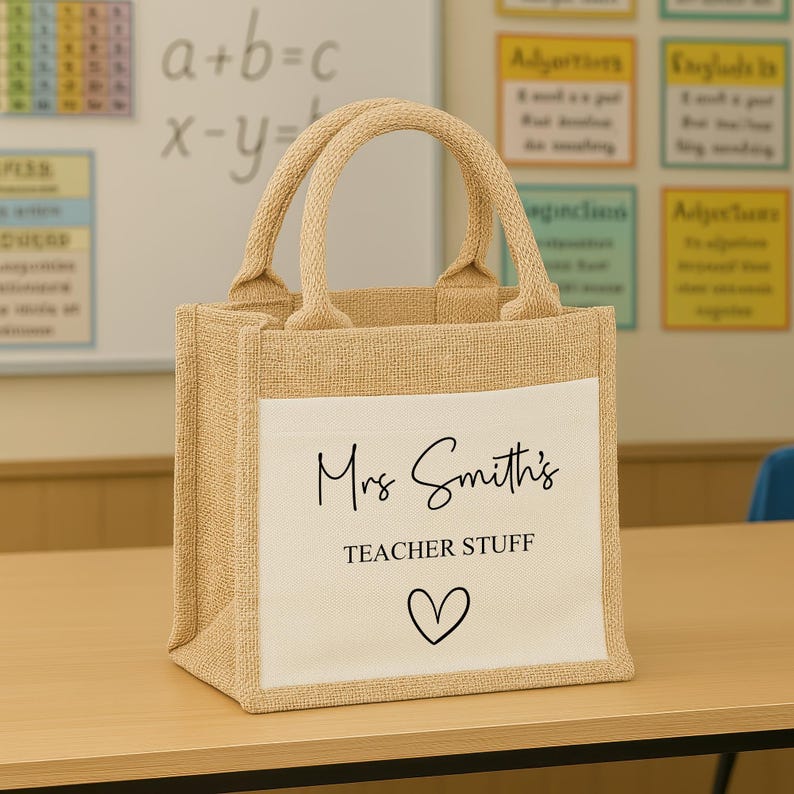 K&ouml;nnte beinhalten: Beige Jute-Tragetasche mit wei&szlig;er Fronttasche. Die Tasche tr&auml;gt den Text "Mrs Smith's TEACHER STUFF" und ein schwarzes Herzsymbol. Die Tasche hat zwei beige Griffe und steht auf einer Holzoberfl&auml;che.