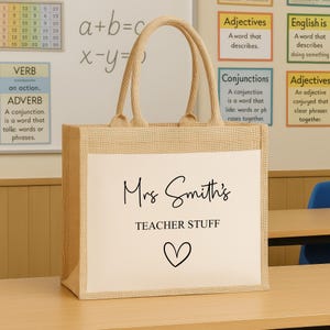 K&ouml;nnte beinhalten: Eine beigefarbene Jutetasche mit wei&szlig;er Frontplatte. Der Text "Mrs Smith's TEACHER STUFF" ist in schwarzer Schrift und Blockbuchstaben gedruckt, darunter ein Herzsymbol. Die Tasche hat beige Griffe.