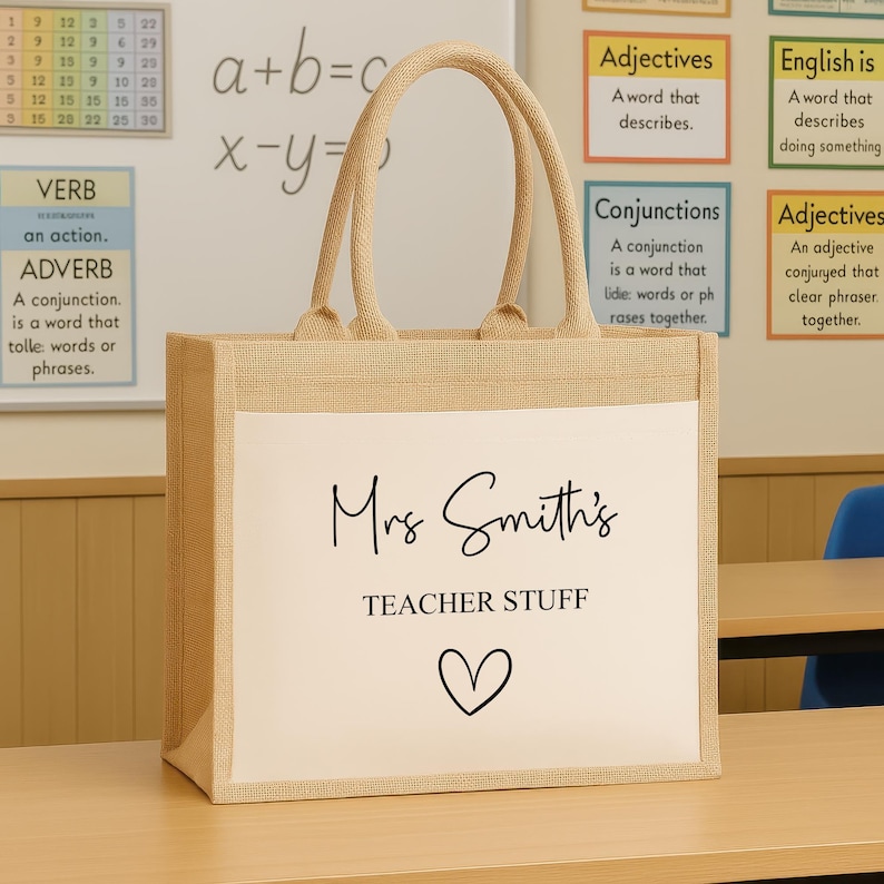 K&ouml;nnte beinhalten: Eine beigefarbene Jutetasche mit wei&szlig;er Frontplatte. Der Text "Mrs Smith's TEACHER STUFF" ist in schwarzer Schrift und Blockbuchstaben gedruckt, darunter ein Herzsymbol. Die Tasche hat beige Griffe.