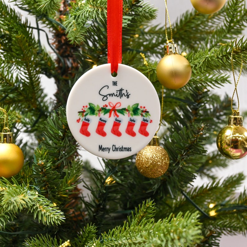 May include: White ceramic Christmas ornament with a red ribbon. The ornament features a red and white illustration of stockings with the names "Daddy", "Mommy", "George", "Sophia", and "Sophia" written on them. The text "The Smith's" and "Merry Christmas" are also printed on the ornament.