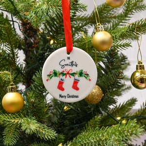 May include: White ceramic Christmas ornament with a red ribbon. The ornament has a green and red holly design with two red stockings hanging from a bow. The text "The Smith's" is written in black cursive script. The text "Mark" and "Francesca" are written in black on the stockings. The text "Merry Christmas" is written in black at the bottom of the ornament.