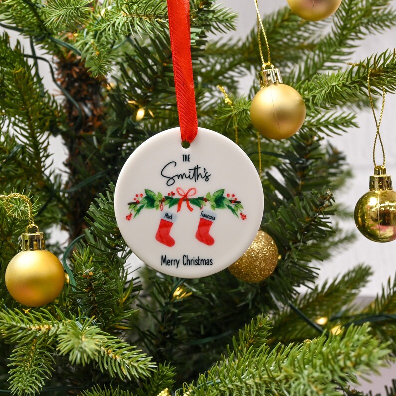 May include: White ceramic Christmas ornament with a red ribbon. The ornament has a green and red holly design with two red stockings hanging from a bow. The text "The Smith's" is written in black cursive script. The text "Mark" and "Francesca" are written in black on the stockings. The text "Merry Christmas" is written in black at the bottom of the ornament.