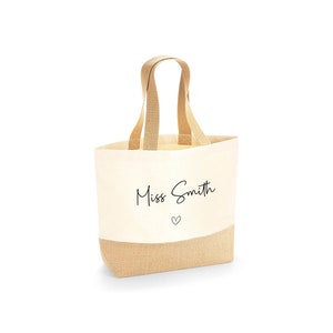 K&ouml;nnte beinhalten: Cremefarbene Tragetasche mit einem Boden und Griffen aus nat&uuml;rlichem Jute. Die Tasche tr&auml;gt den Text "Miss Smith" in einer handschriftlichen Schriftart, mit einem kleinen Herz darunter. Zum Tragen von Gegenst&auml;nden.