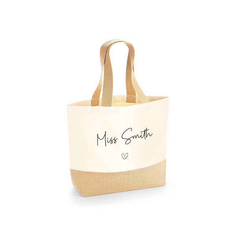 K&ouml;nnte beinhalten: Cremefarbene Tragetasche mit einem Boden und Griffen aus nat&uuml;rlichem Jute. Die Tasche tr&auml;gt den Text "Miss Smith" in einer handschriftlichen Schriftart, mit einem kleinen Herz darunter. Zum Tragen von Gegenst&auml;nden.