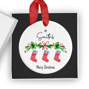 May include: White ceramic Christmas ornament with a red ribbon, green holly, and three red stockings with the names "Daddy", "Mummy", and "Sophia" hanging from a green garland. The text "The Smith's" and "Merry Christmas" are printed on the ornament.