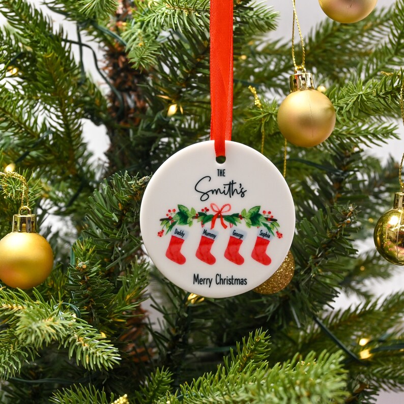 May include: White ceramic Christmas ornament with a red ribbon. The ornament features a green holly wreath with four red stockings hanging from it. The stockings are labeled "Daddy", "Mommy", "George", and "Sophia". The text "The Smith's" and "Merry Christmas" are printed on the ornament.