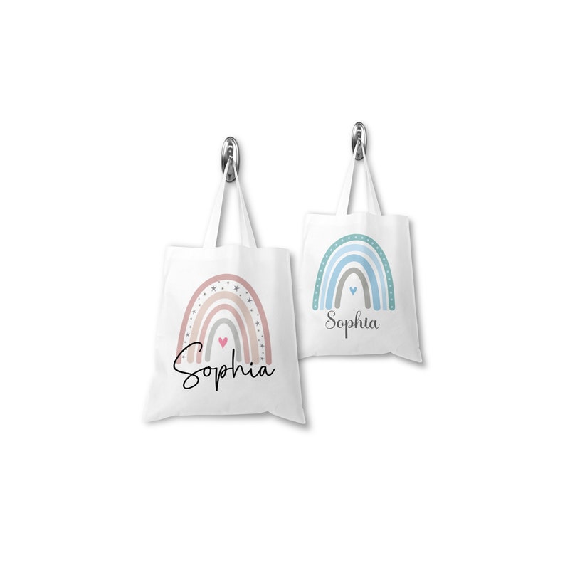 K&ouml;nnte beinhalten: Zwei wei&szlig;e Canvas-Tragetaschen mit Regenbogen-Designs und dem Namen "Sophia" auf jeder Tasche. Ein Regenbogen ist in Rosa- und Pfirsicht&ouml;nen gehalten, der andere in Blau- und Gr&uuml;nt&ouml;nen.