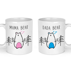May include: Two white ceramic mugs with black text and illustrations. The mug on the left says "Mama Bear" and features a white bear with pink accents. The mug on the right says "Dada Bear" and features a white bear with blue accents.